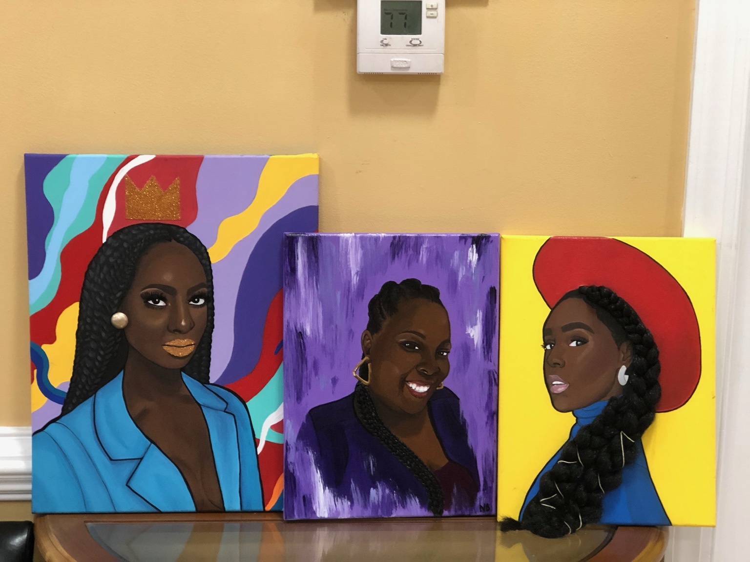 Three mixed-media portraits by Nika B displayed together, featuring a gold crown, a purple background, and a red hat